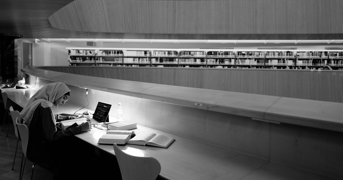 A student studying at a desk lit by a lamp in a quiet university library, books open in front of them