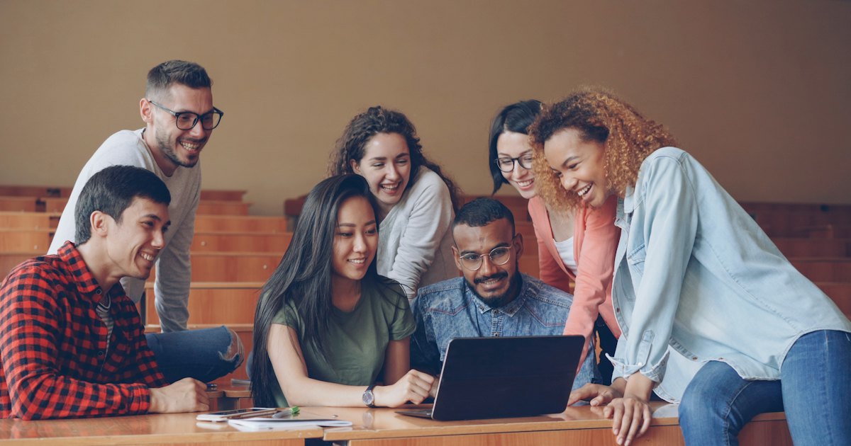 A group of university students gathered around a laptop in a lecture hall, talking and smiling