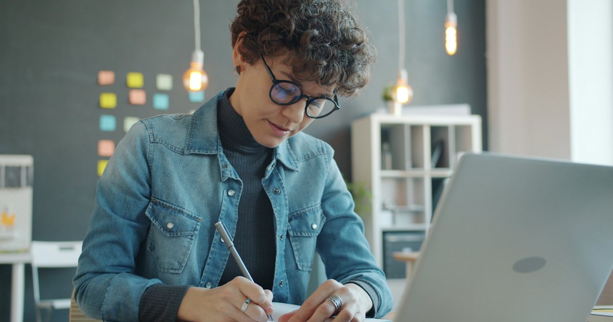 A writer with glasses taking notes in a notebook beside an open laptop