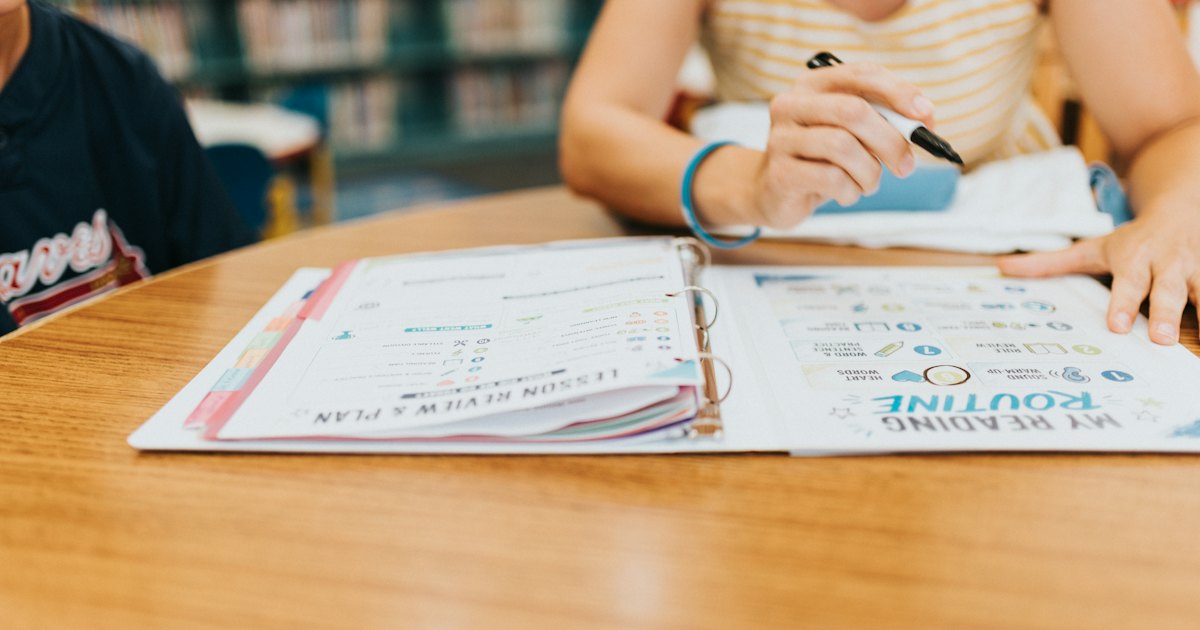 A student writing in an open binder on a wooden table, with reading planning sheets visible