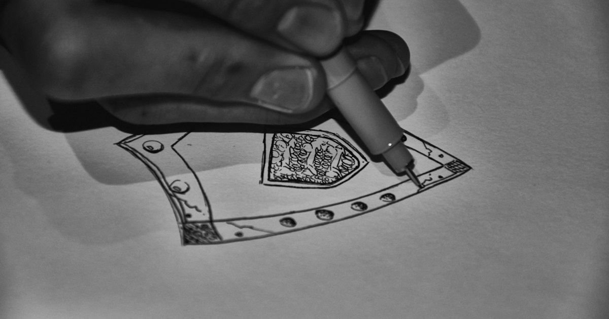 A hand drawing a detailed emblem design with a fine-tip pen on paper