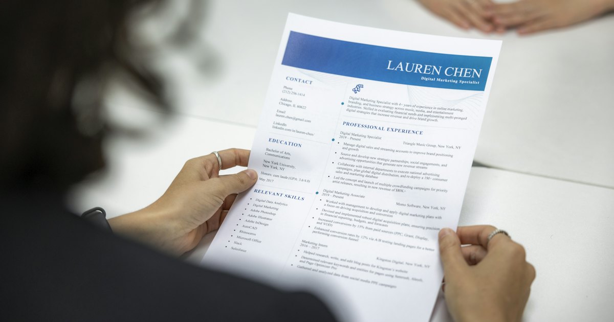 A person reviewing a printed resume at a desk, with another person's hands visible across the table