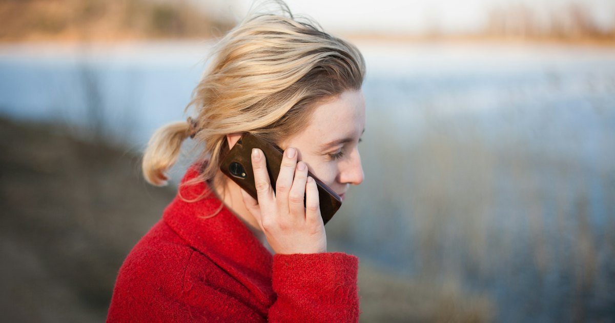 A woman in a red coat speaking on her phone outdoors beside a calm lake at sunset