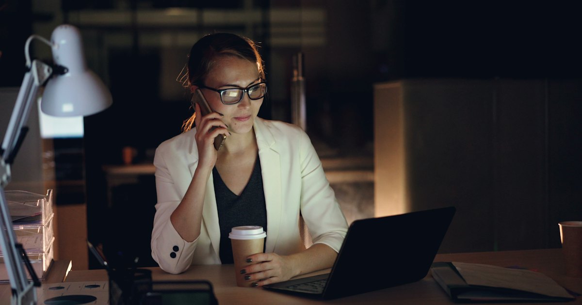 A woman working late at a desk with a phone in one hand and a laptop open in front of her