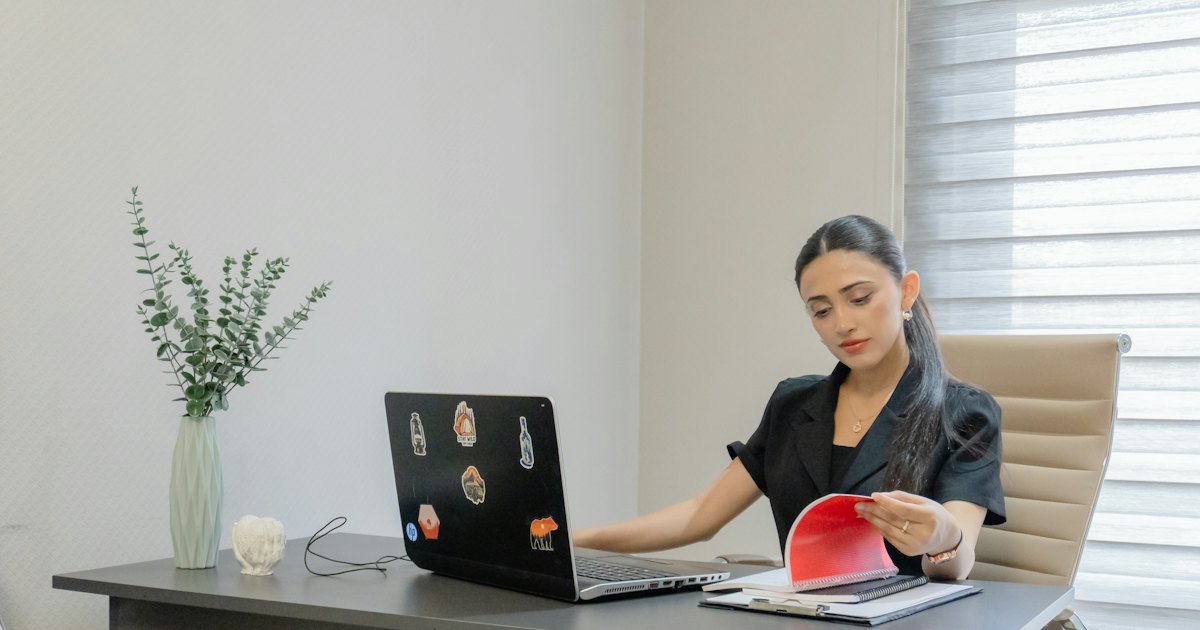 A woman at a desk reading printed pages beside an open laptop