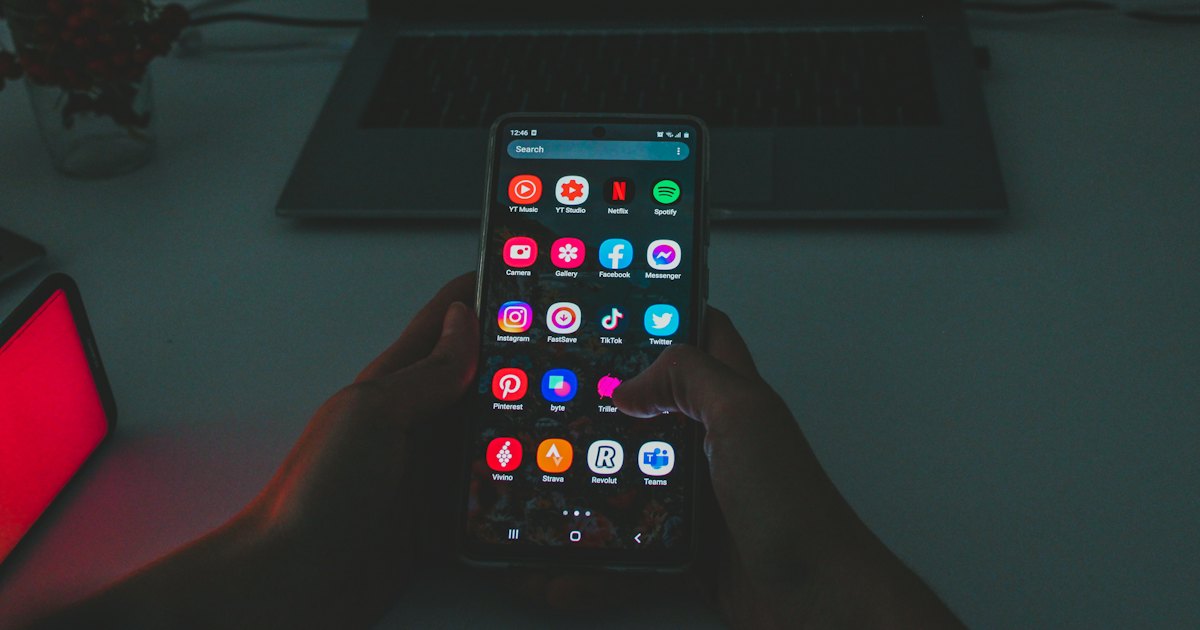 A hand holding a smartphone with a home screen full of app icons, a laptop in the background