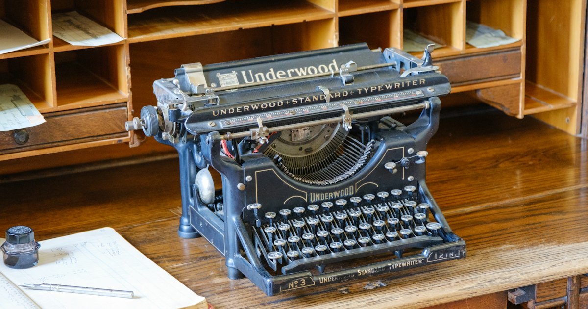 A vintage Underwood typewriter sitting on a wooden desk beside an open ledger and an inkwell