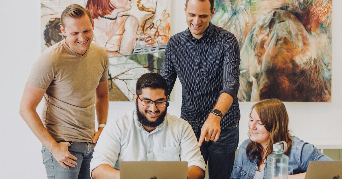 Four coworkers smiling around a laptop at an office table