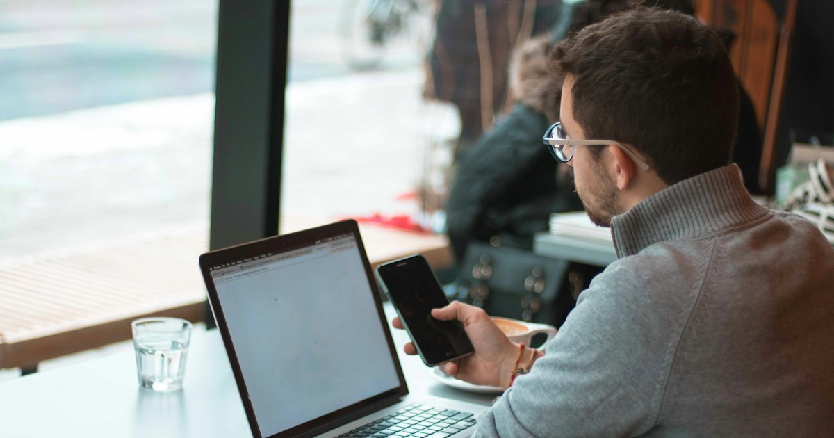 A man at a cafe table by the window with a laptop open and a phone in his hand