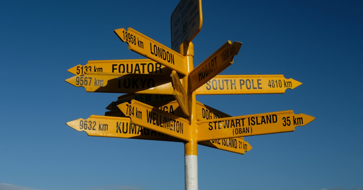 A yellow signpost crowded with arrows pointing to far-off places, set against a bright blue sky