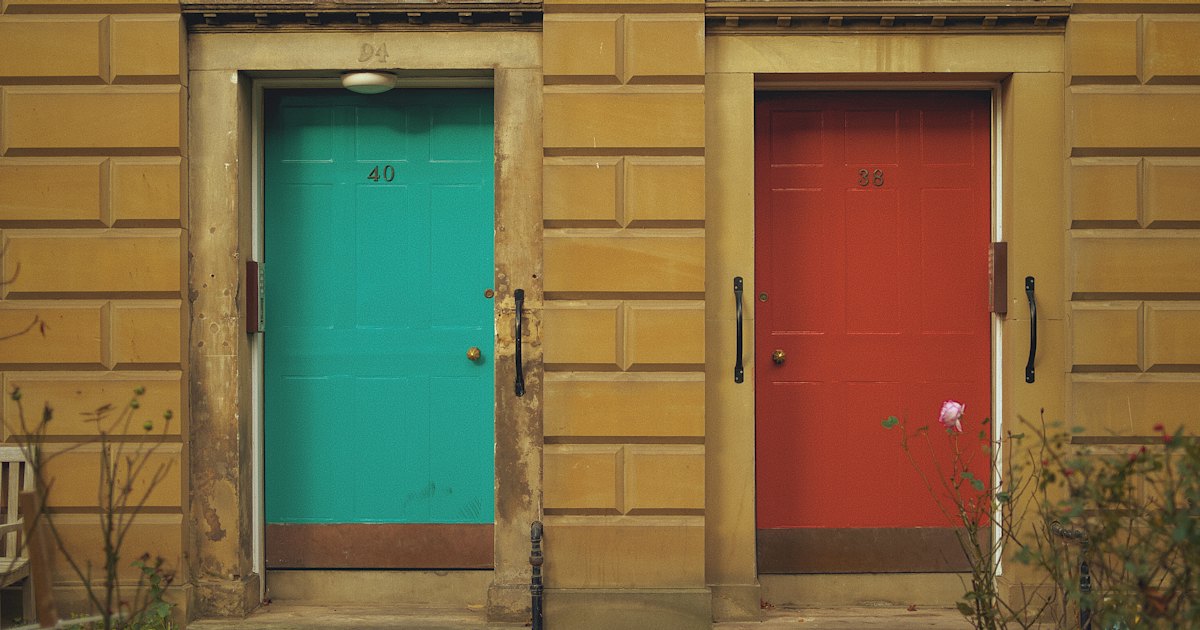 Two painted front doors side by side on a building, one teal and one red, with a rose bush beneath the red door