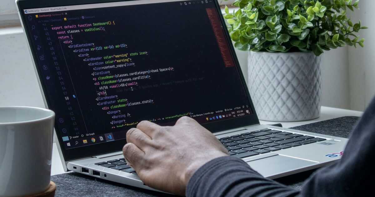 A person typing code on a laptop at a home workspace with a plant and mug nearby