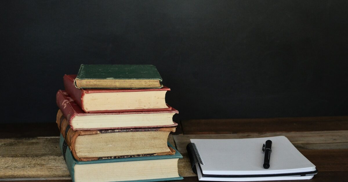 A tall pile of books beside paper and a ballpoint pen on a desk