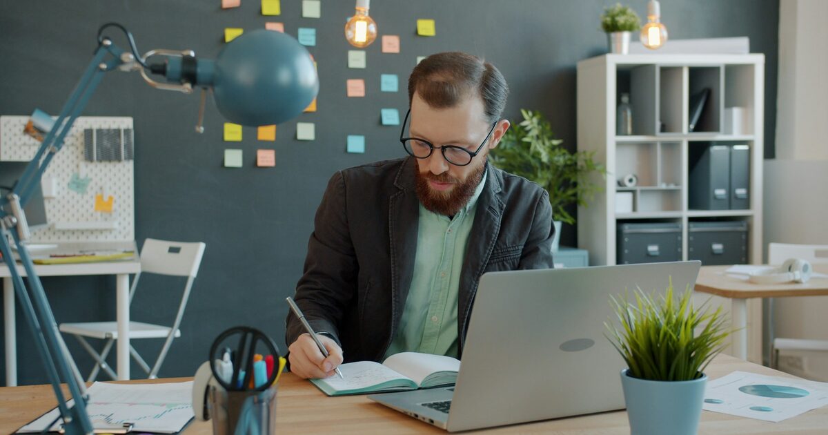 A man writing in a notebook beside an open laptop at his desk